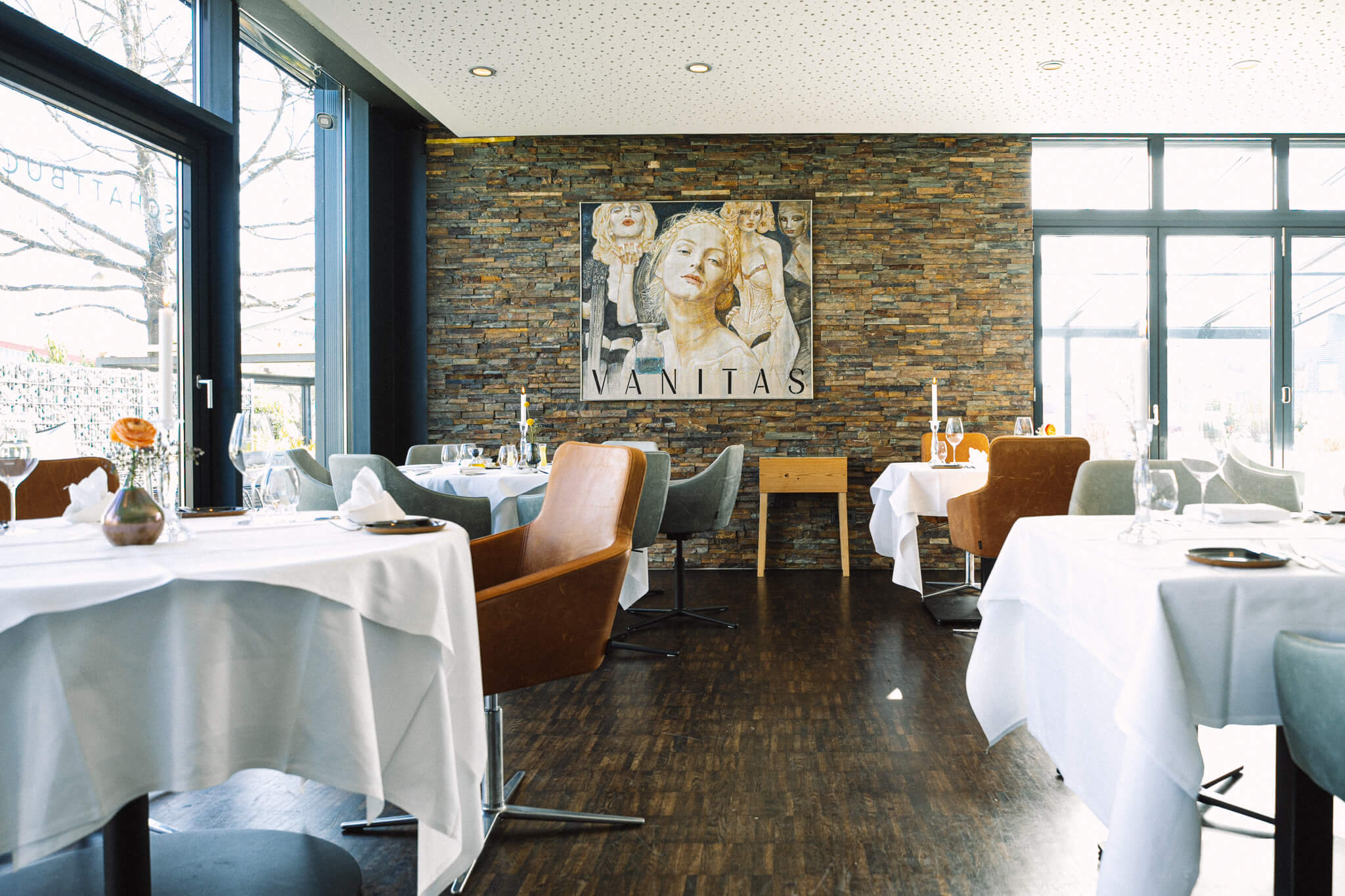 Mehrere runde und rechteckige Tische, weiß eindeckt, in einem hellen Raum mit dunklem Parkettboden und einer Steinwand, die ein Gemälde schmückt