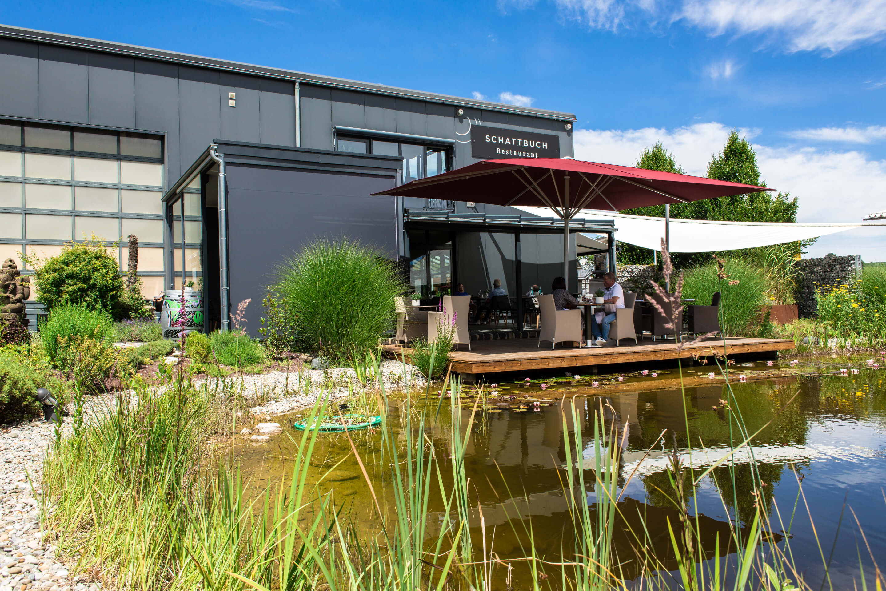 Außenaufnahme des Kräutergartens mit Teich und einem aufgespannten Sonnenschirm sowie Kubus