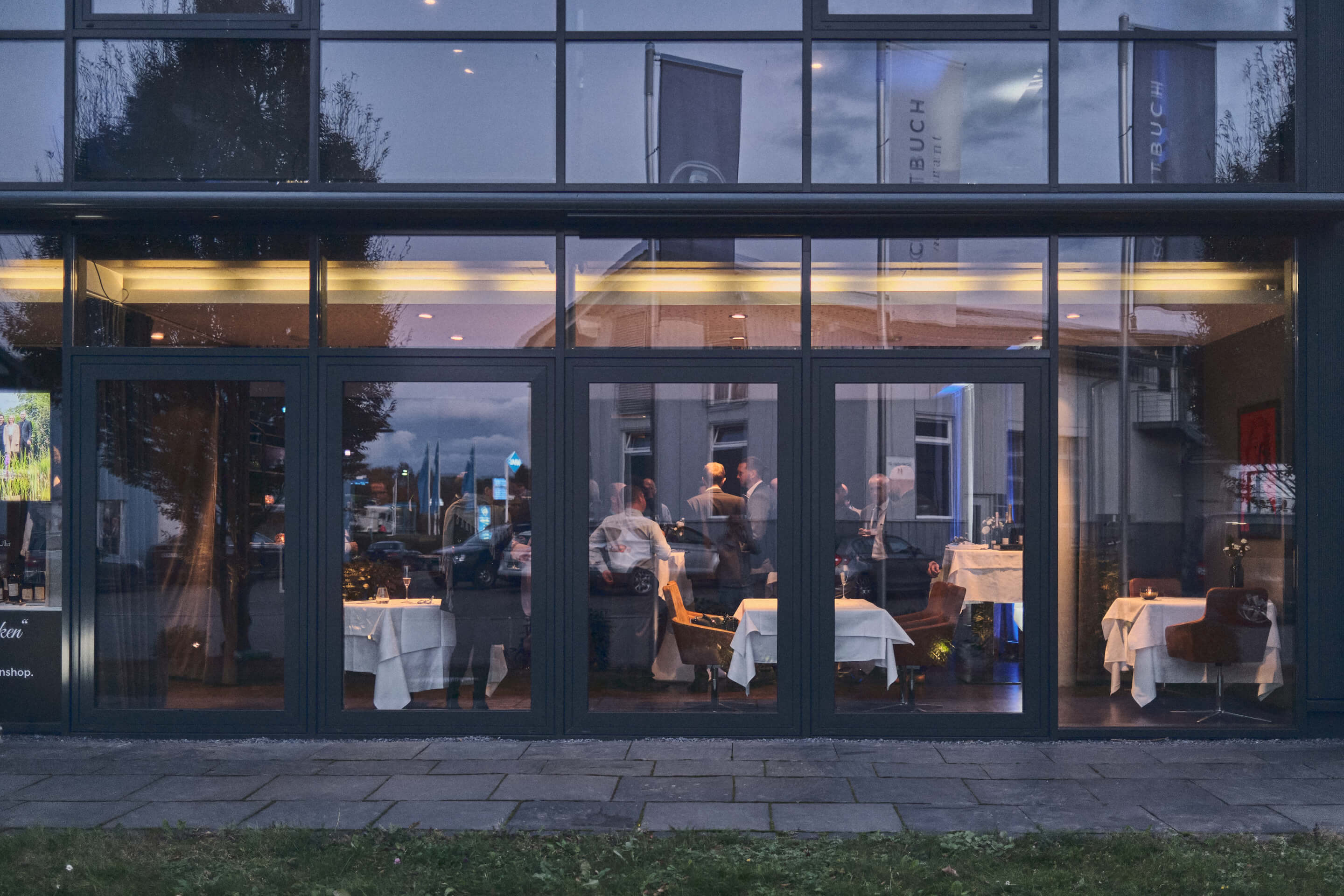 Außenaufnahme der Glasfront mit Menschen, die zusammenstehen und bei einem Aperitif sich an Stehtischen unterhalten