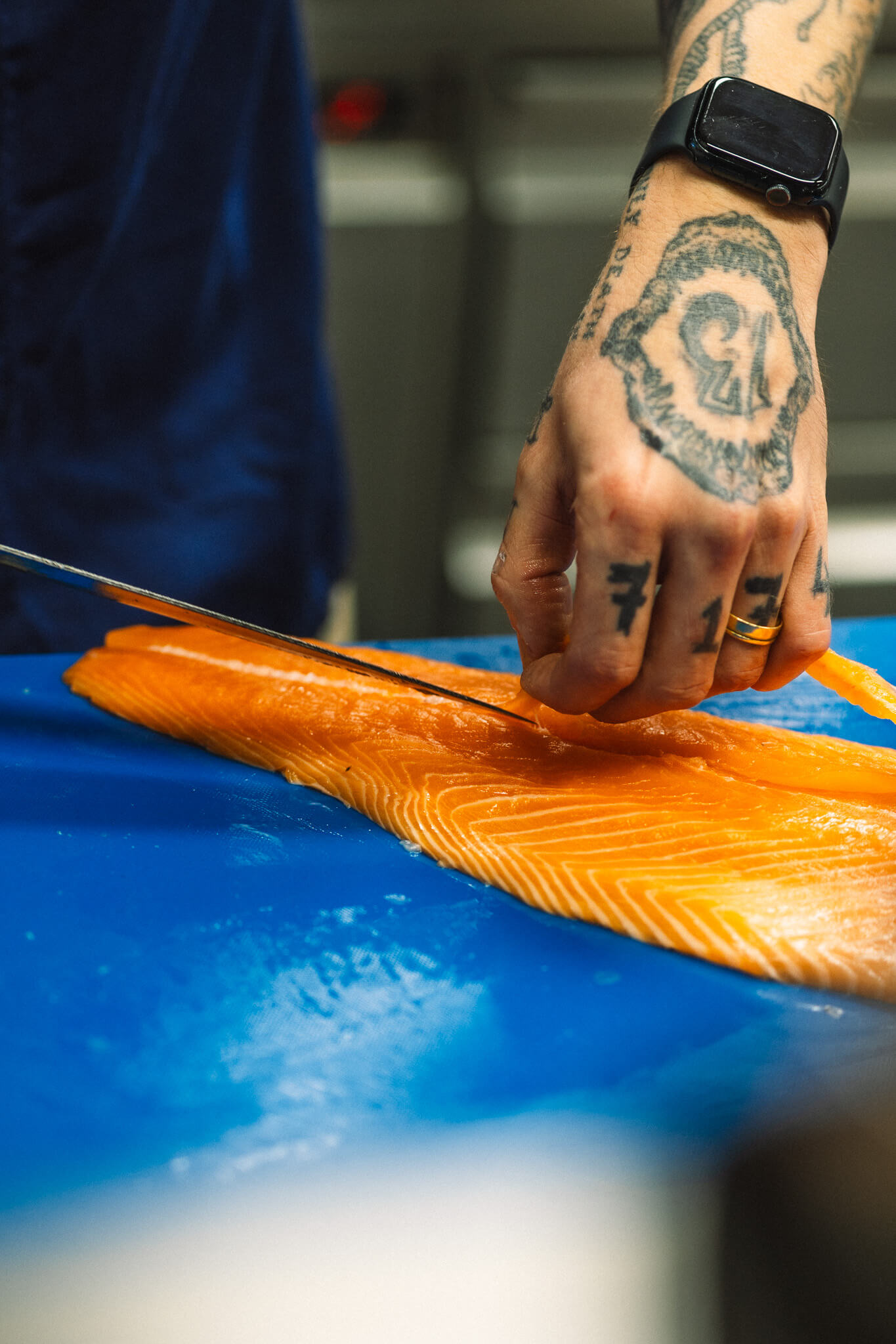 Eine tätowierte Hand hat ein Messer in der Hand und filetiert damit einen rohen Fisch