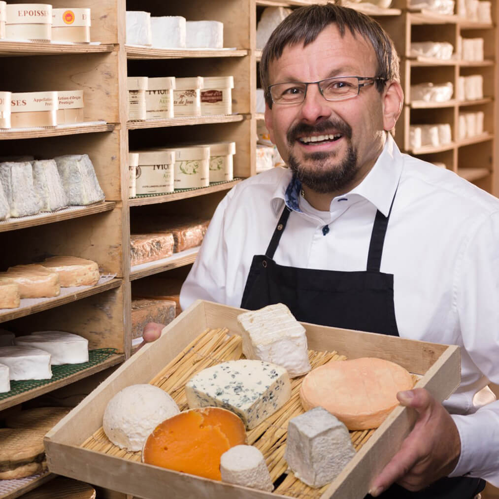 Mann im weißen Hemd und mit Schürze hält ein Brett mit verschiedenen Weichkäsen in der Hand im Käselager