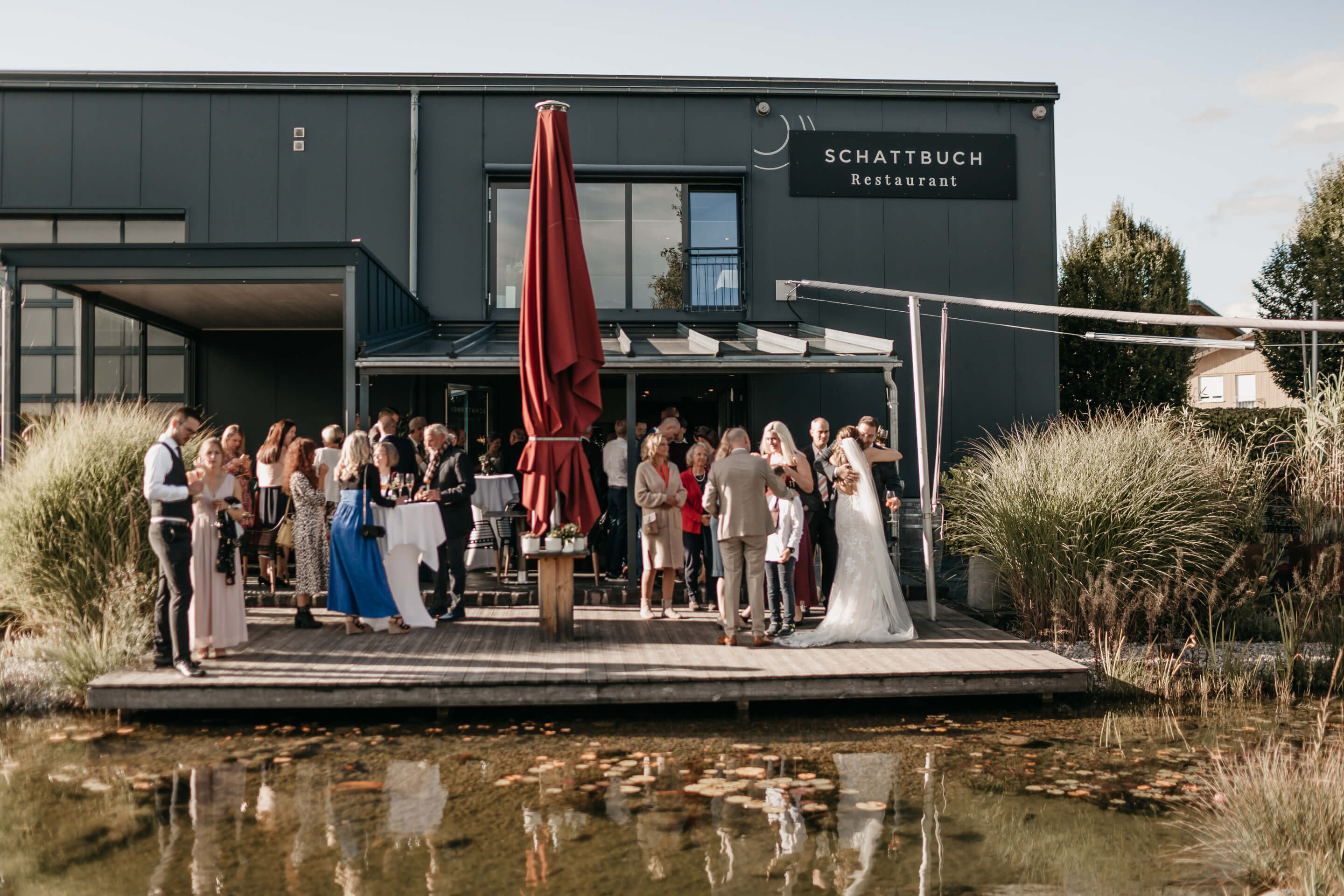 Eine Gruppe von Menschen, die Teil der Hochzeitsgesellschaft sind, stehen auf einer Terrasse am Teich