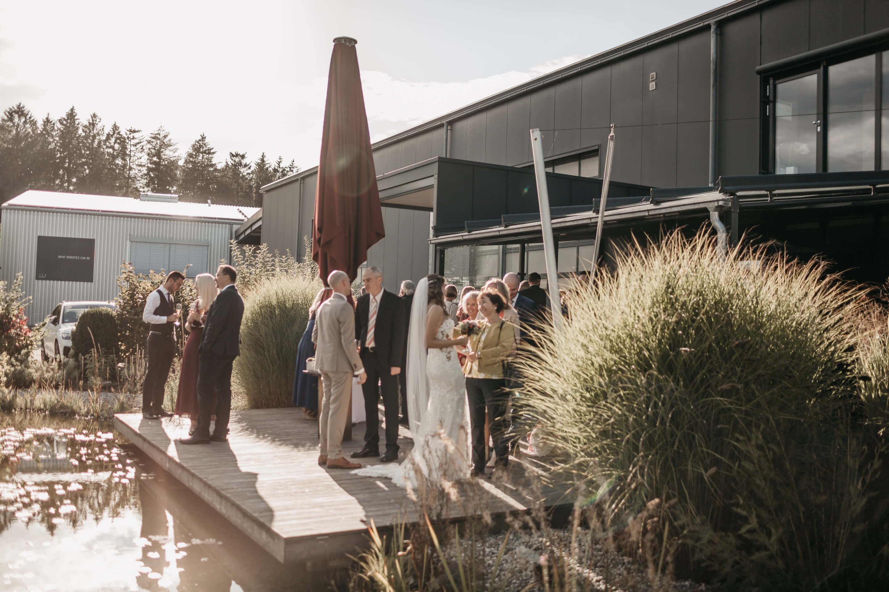 Eine Gruppe von Menschen, die Teil der Hochzeitsgesellschaft sind, stehen auf einer Terrasse am Teich mit der Braut