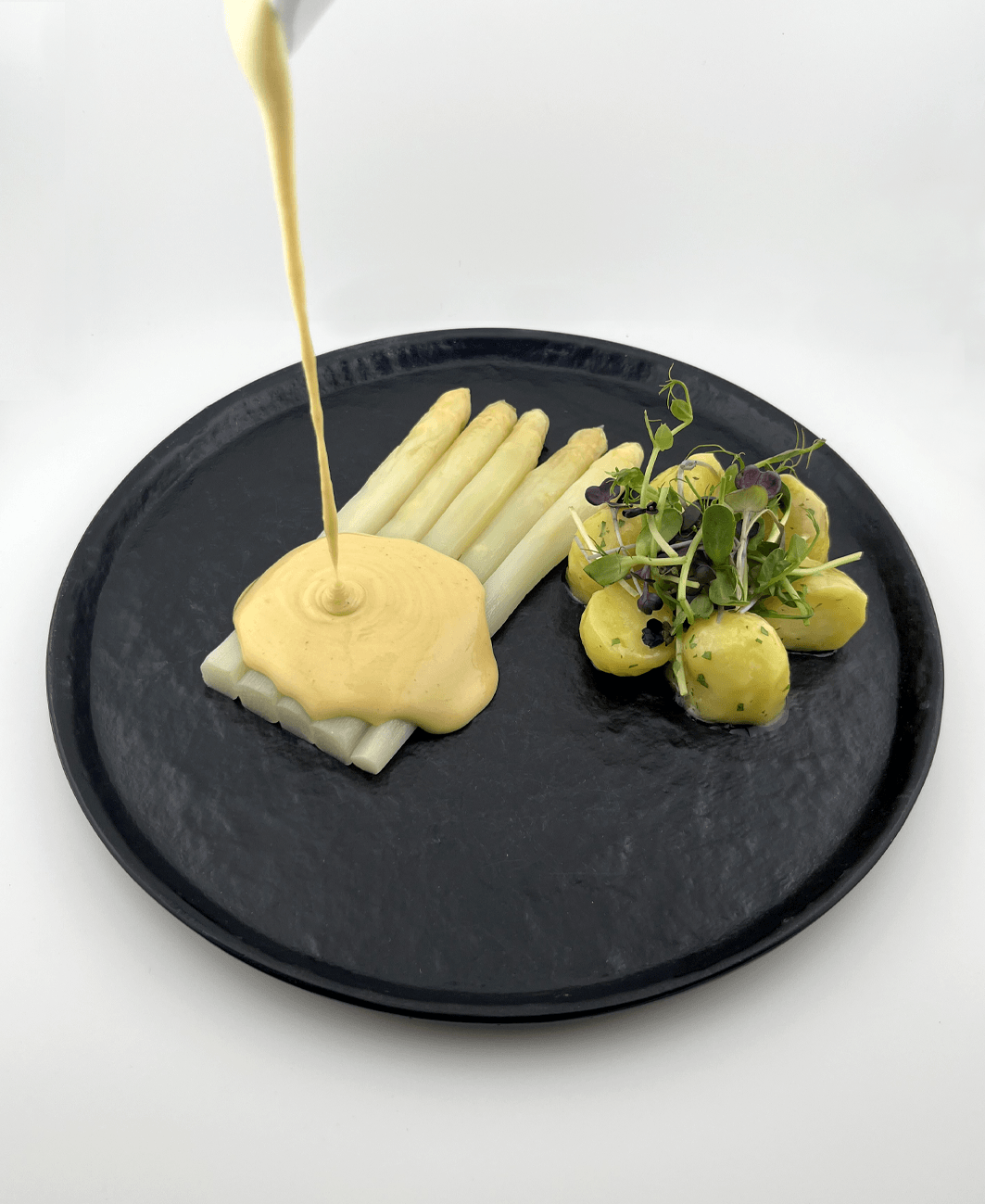Dunkler Teller mit weißem Spargel, über diesen eine Sauce Hollandaise gegeossen wird, dazu Kartoffeln