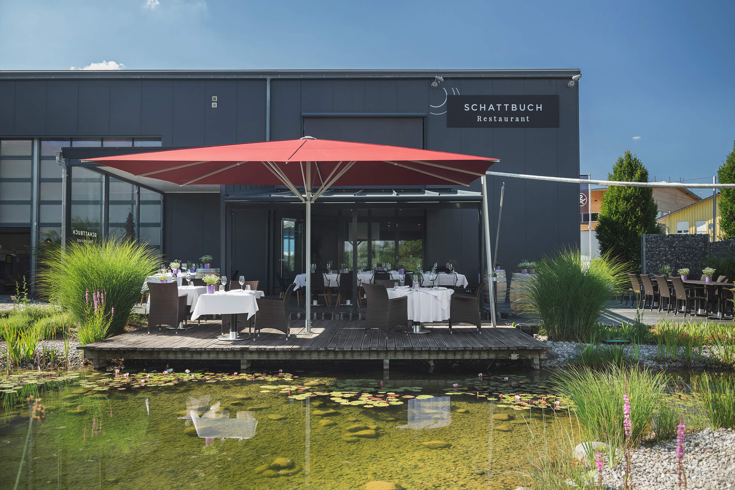 Teich mit Terrasse unter einem großen roten Sonnenschirm, auf der auch eingedeckte Tische Platz finden