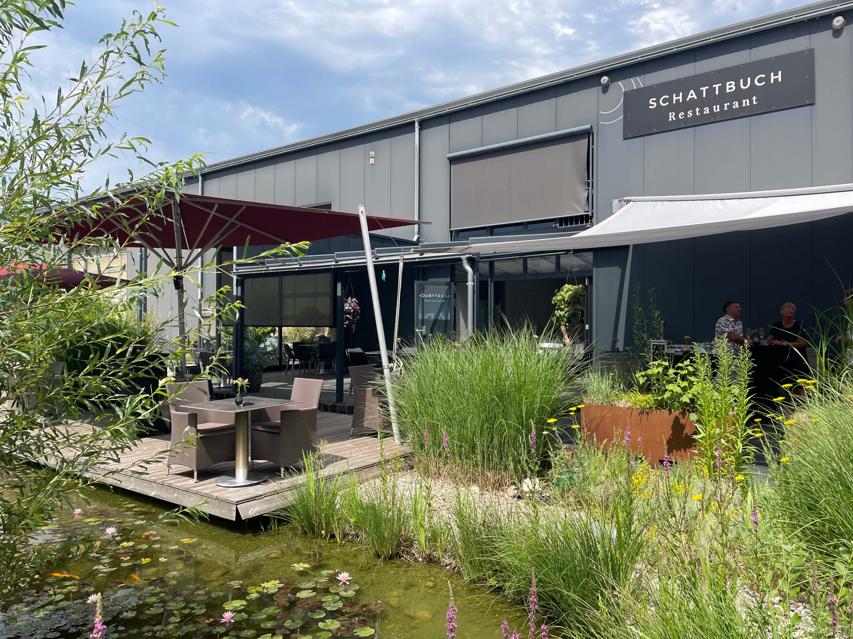 Eine Terrasse mit Holzdielen und mehreren Tischen an einem mit Gräsern und Pflanzen zugewachsenen Teich
