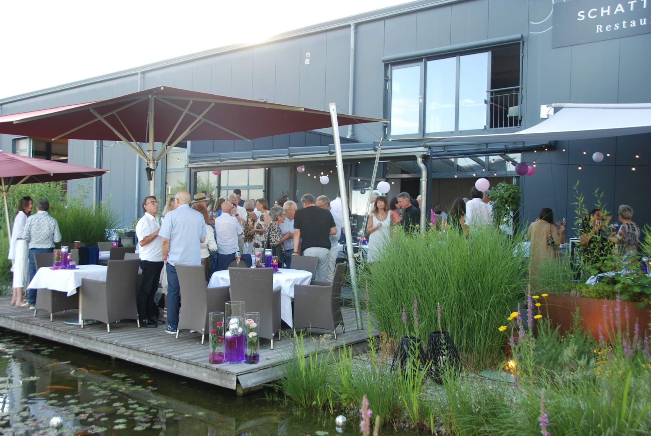 Terrasse am Teich mit grünen Seerosen und Sträuchern, auf der Menschen sich gesellig unterhalten und den Abend einläuten